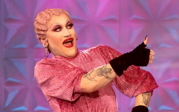 Drag Queen Joe Black in a pink dress, with black fingerless gloves giving a thumbs up sign to someone out of the picture.